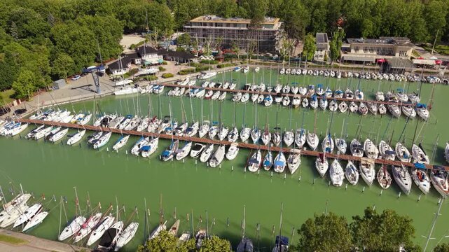 Top-down aerial view of sailboats and yachts moored along a marina dock, with long geometric mast shadows stretching across green water.