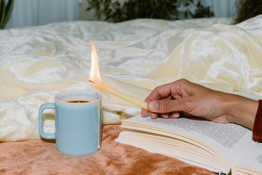 Person lighting a candle over a cup of coffee and an open book in a cozy bedroom setting for relaxation and wellness concepts