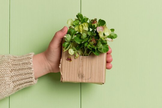 Child holding a handmade cardboard vase with real green flowers and leaves over a mint green wooden surface for nature craft and sustainability concepts