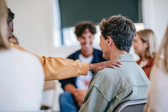 Close up of a hand on a shoulder between two teenagers during a group session, showing compassionate physical touch for peer support and mental health content
