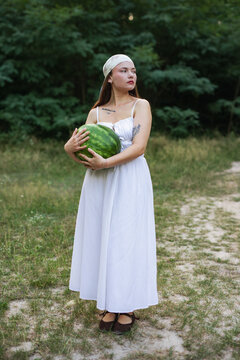 Young woman in white sundress holding large watermelon in nature