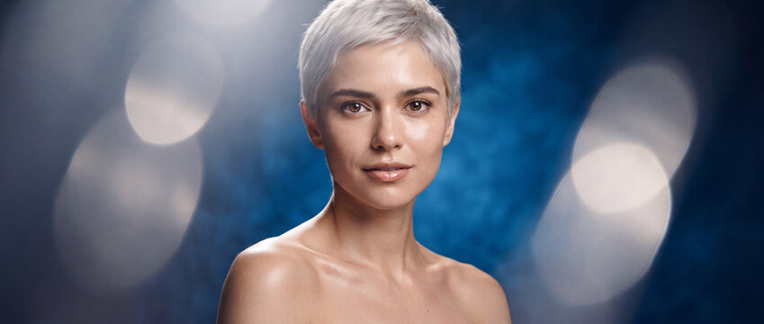 Beauty portrait of a woman with short hair featuring glowing natural skin, silver blond pixie cut, and soft lighting against a blue bokeh background, ideal for skincare and cosmetics
