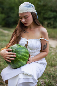 Young woman crouching on green grass and holding large watermelon