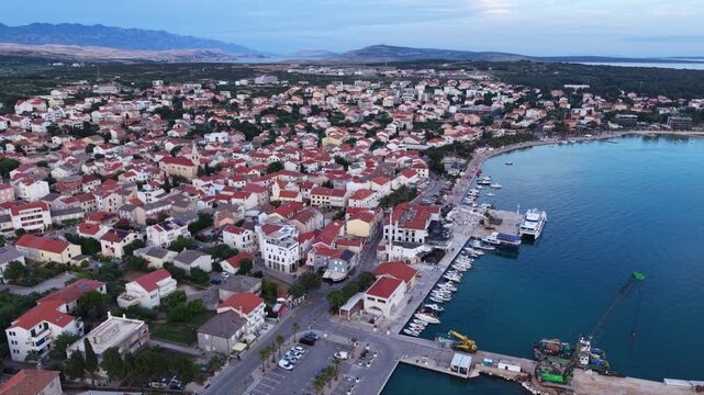 Aerial panoramic view of Novalja, Croatia, showing the coastal town&rsquo;s dense red-roofed architecture, lively central promenade and traditional churches, with the dramatic rocky mountains of Pag Island 