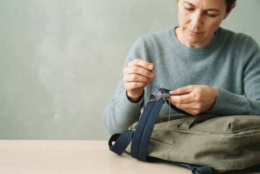Adult repairing a torn backpack strap with a needle and thread against a muted green background for back to school and caregiving content