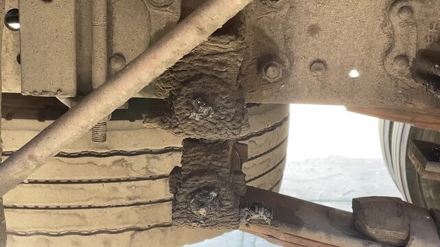 Detailed view beneath a dusty truck showing a tire, axle, suspension parts, grease, dirt, and metal structures, documenting mechanical state during an outdoor heavy vehicle inspection today.