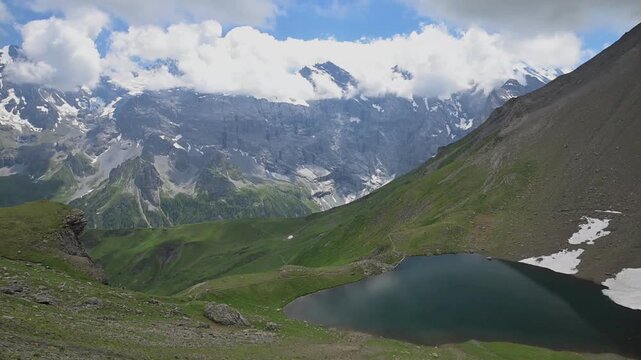 Dark alpine lake surrounded by green slopes, rocky peaks, snowfields and dramatic clouds in the Swiss Alps.