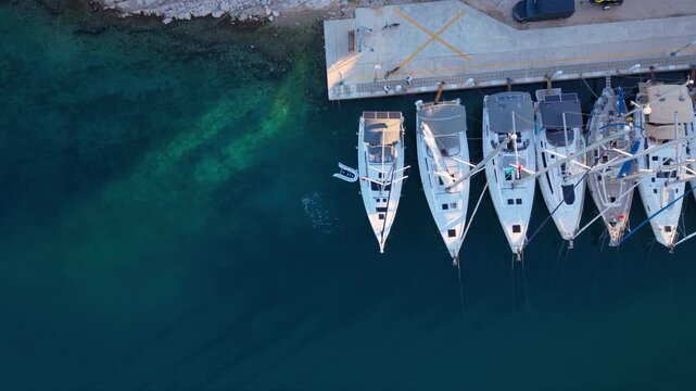 Top-down aerial view of several sailing yachts moored side by side at a marina pier, with their masts creating strong geometric lines above deep blue transparent water near a rocky shoreline.