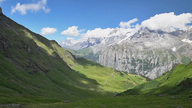 Sweeping view of green mountain slopes, rugged limestone peaks and lingering snow beneath a bright blue summer sky.
