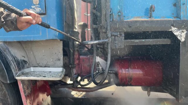 Water sprays across a blue truck body and metal panel, washing away dirt and grime during vehicle cleaning, with droplets and runoff visible on the exterior surface outdoors during service.