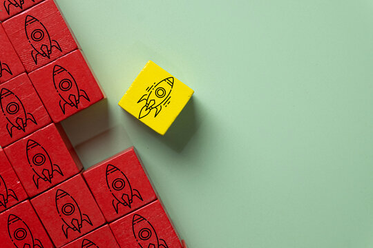 Standing out from the crowd and leadership concept. A yellow wooden block with a launching rocket separating from red blocks with stationary rockets on a green background with copy space.