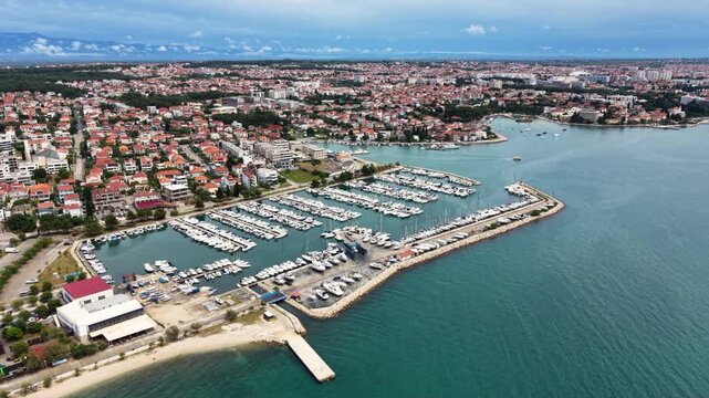 High aerial view of Luka Sukosan marina on the Croatian Adriatic coast, with long breakwaters, rows of moored yachts and sailboats, a coastal town and forested hills extending along the shoreline. 