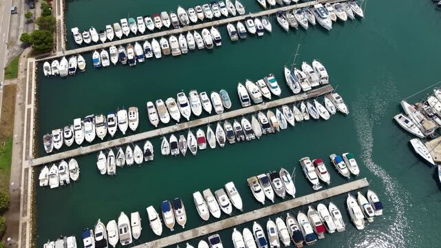 Aerial view of a large Mediterranean marina packed with yachts and sailboats, with long stone breakwaters, multiple piers and dense rows of moored vessels beside a coastal residential area.
