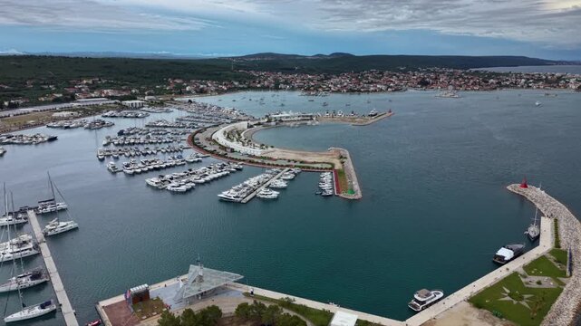 High aerial view of a large Mediterranean marina filled with rows of yachts and sailboats, protected by a curved breakwater beside a coastal town and waterfront promenade on the Adriatic coast.