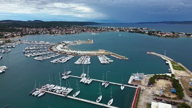 High aerial view of a large Mediterranean marina filled with rows of yachts and sailboats, protected by a curved breakwater beside a coastal town and waterfront promenade on the Adriatic coast.