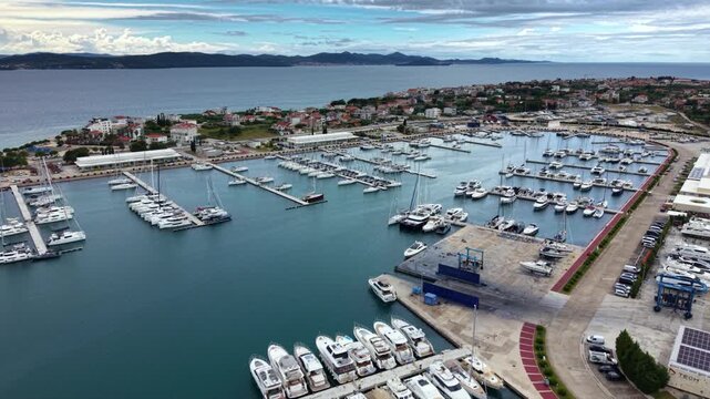 High aerial view of Luka Sukosan marina on the Croatian Adriatic coast, with long breakwaters, rows of moored yachts and sailboats, a coastal town and forested hills extending along the shoreline. 