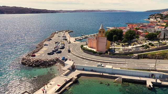 Top-down aerial view of a small historic church in Karlobag, Croatia, beside a curved coastal road with several motorcycles riding past, surrounded by parking areas and traditional red-roofed building