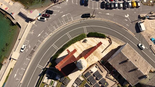 Top-down aerial view of a small historic church in Karlobag, Croatia, beside a curved coastal road with several motorcycles riding past, surrounded by parking areas and traditional red-roofed building