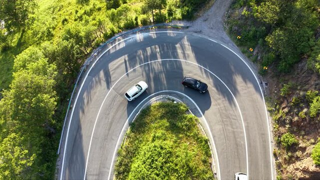Top-down aerial drone view of a semi truck negotiating a tight hairpin bend on a mountain road surrounded by dense green forest, with cars passing through the sharp curve.
