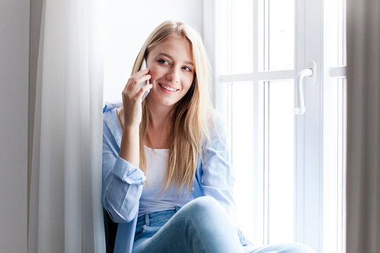 Young woman talking on mobile phone at home by window. Happy girl holding cell phone and calling, enjoying conversation. People connection, communication. Lifestyle moment, everyday life