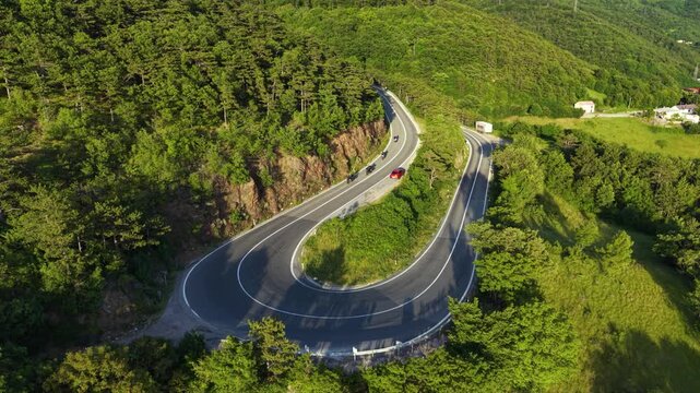 Aerial drone view of motorcyclists riding around a dramatic hairpin bend on a winding mountain road surrounded by dense green forest, with cars and a truck traveling nearby.