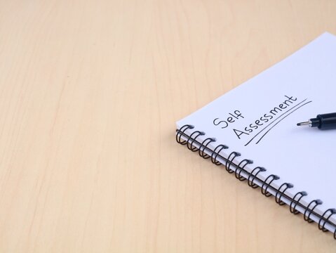 Self assessment handwritten on book on wooden desk with pen and copy space. Concept of personal evaluation, performance review, self improvement, business planning, and career development