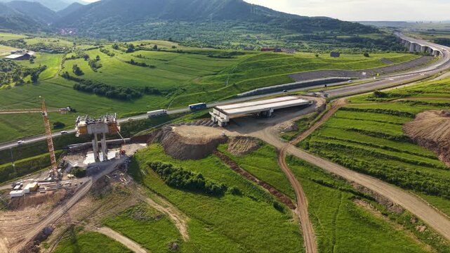 Aerial drone view of highway bridge construction across green rural fields. Concrete piers, cranes, earthworks and unfinished overpass sections show major transport infrastructure development project.
