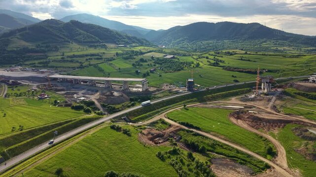 Aerial drone view of highway bridge construction across green rural fields. Concrete piers, cranes, earthworks and unfinished overpass sections show major transport infrastructure development project.