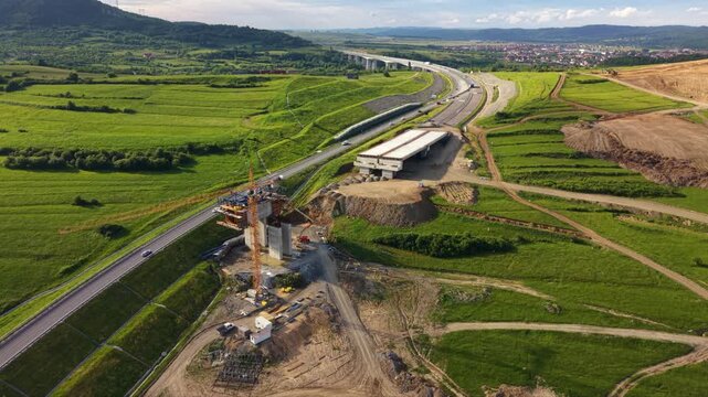 Aerial drone view of highway bridge construction across green rural fields. Concrete piers, cranes, earthworks and unfinished overpass sections show major transport infrastructure development project.