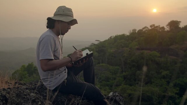 Man sitting on a hill writing in a journal with the warm orange afternoon sun in the background, capturing mindfulness, reflection, and peaceful moments in nature.
