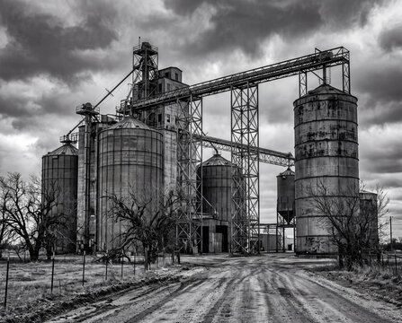 Dramatic black and white landscape of a rural agricultural grain elevator and silos under a moody cloudy sky