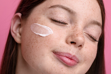 Beautiful girl with freckles and cream on her face against pink background, closeup