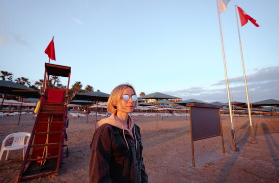A young woman in a denim jacket and reflective sunglasses stands on an empty beach at sunset by a lifeguard chair and rows of closed umbrellas, evoking calm, travel, and quiet contemplation.