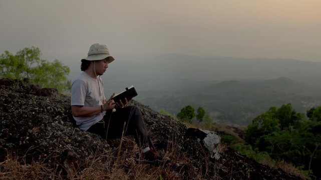 Man enjoying a book on a hilltop during sunset, with mountains and beautiful natural scenery in the background