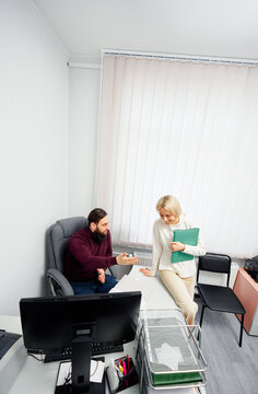 A bearded man in a burgundy sweater chats with a blonde colleague who holds a green folder in a bright, modern office. They discuss work near a desk with a computer.