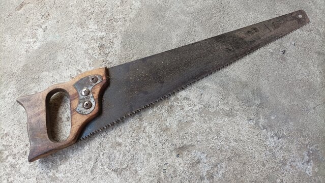 Old rusty hand saw with a wooden handle lying on a concrete surface.