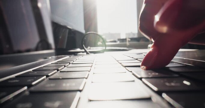 Low angle closeup hands typing on laptop keyboard at desk by window in office during morning light, tracking shot slow motion.