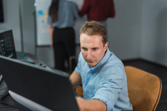 Man developer studies his monitor while reviewing AI output for errors, code visible on a second screen and colleagues planning at a whiteboard behind him.