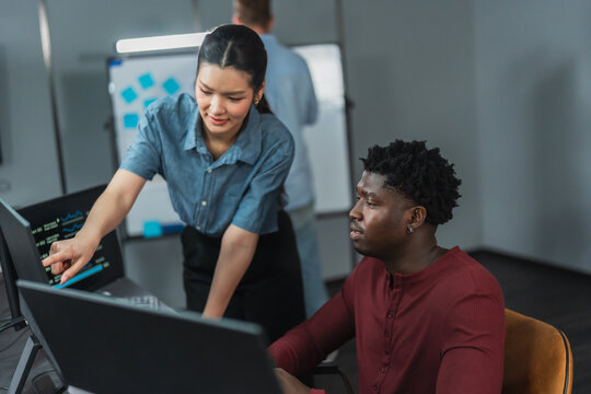 Woman developer points at a monitor to show a colleague how to work with AI coding tools, demonstrating how the workflow speeds up software development in a tech office.
