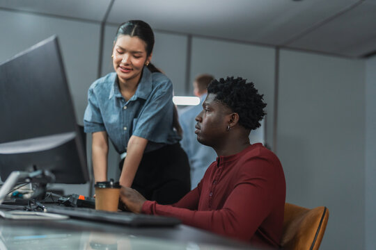 Female developer stands beside a seated developer at a workstation with coffee and monitors, discussing work in a modern technology office.