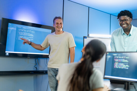 Smiling man points at code displayed on a wall screen while a colleague laughs during a team session in a blue-lit tech office.