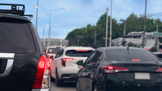 Rear view of multiple cars waiting in a heavy traffic jam on an urban highway. Highlighting red tail lights during a busy daytime commute. Background of green forest under blue sky and white clouds.