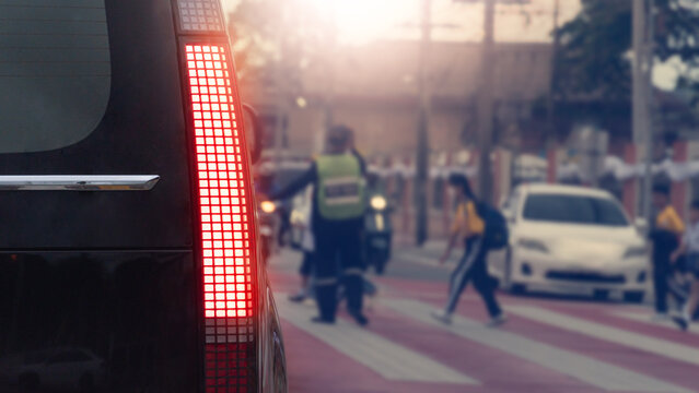 Rear side of car stopped near a pedestrian crossing on a city street. Showcasing a bright vertical tail light and urban traffic under a bright daytime sun. With blurry image of students passing by.