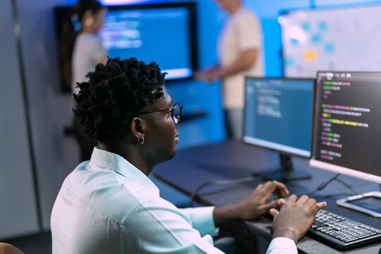Man in glasses types at a workstation with two screens of code while colleagues talk near a whiteboard in a blue-lit tech office.