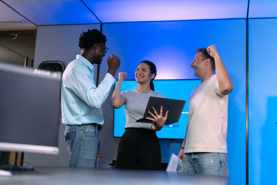 Three colleagues raise their fists and smile around a laptop as they celebrate a project win in a blue-lit technology office.