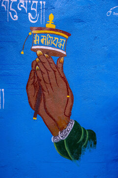 A hand holding a Tibetan prayer wheel painted on a blue wall. The mantra &ldquo;Om Mani Padme Hum&rdquo; is inscribed on the wheel. Dharamsala, Himachal Pradesh, India.