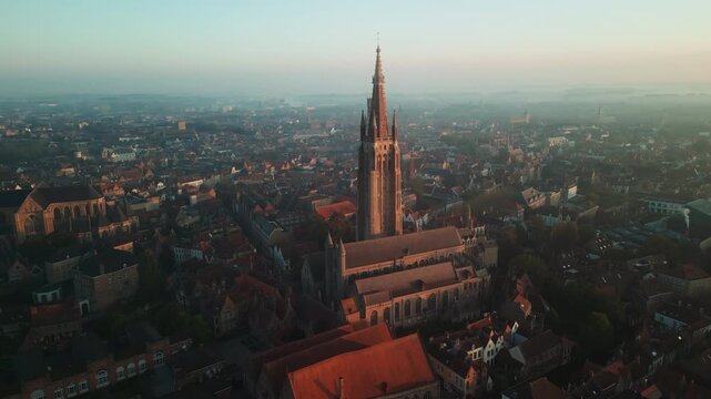 Morning drone view frames the Church of Our Lady above dense red roofs in Bruges, Belgium, with the Belfry and historic skyline fading into soft haze.