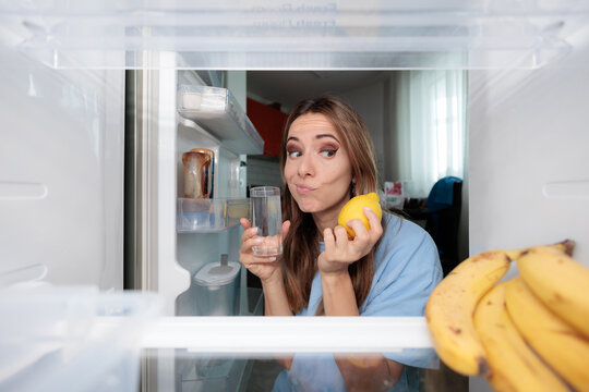 Puzzled Young Woman Holding Lemon And Glass Water Near Refrigerator. Confused female doubts internet health hack while looking inside open kitchen fridge