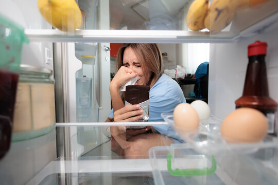 Sad Crying Woman Eating Chocolate Bar Inside Open Kitchen Refrigerator. Emotional female weeps while consuming sweet snacks near open fridge at night