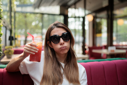 Bored Young Woman Wearing Sunglasses Holding Cold Colorful Cocktail Drink. Tired female makes funny facial expression while waiting inside terrace cafe